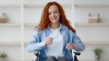 Kızıl saçlı genç bir kız evinde tekerlekli sandalyesinde oturuyor ve gülümsüyor. Arka planda basit bir dekor var. Neşeli tavırlarını ve rahatlığını vurguluyor..