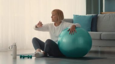 Emekli beyaz bir kadın evinde büyük mavi bir egzersiz topu kullanarak egzersiz yapıyor. Modern yaşam alanındaki günlük rutinine uyum sağlayarak odaklanmış görünüyor..