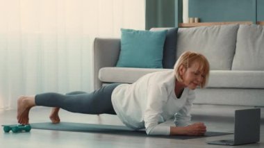 Emekli bir kadın evinde egzersiz yapıyor, güç ve dengeye odaklanıyor. Bir paspasın üzerinde egzersiz yapıyor, dijital bir rehberi takip ediyor, aktif bir yaşlı hayatı sergiliyor..