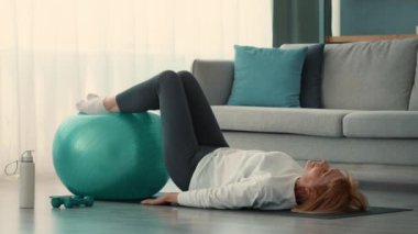 Son sınıf öğrencisi bir kadın evde egzersiz yapıyor. Spor yaparken egzersiz topunda denge kuruyor. Sağlıklı yaşam için odaklanmış bir ifade sürdürüyor..