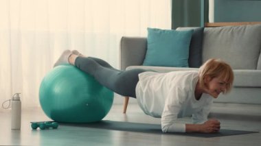 Yaşlı bir kadın oturma odasında dengeli bir top kullanarak egzersiz yapıyor. Dengesini ve gücünü korumaya odaklanıyor. Emekliliğine olan bağlılığını gösteriyor..