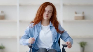 Kıvırcık kızıl saçlı genç bir kız evdeki tekerlekli sandalyesinde güven içinde oturuyor. Nötr renkli duvarları ve rafları olan iyi ışıklandırılmış bir bölgede rahatlamak için hoş bir ortam yaratıyor..
