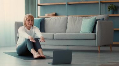 Üst sınıftan bir kadın laptoptaki egzersizleri takiben evde egzersiz yapıyor. Oturma odasında, rahat bir kanepe ve basit bir dekorun bulunduğu bir paspasın üzerinde oturuyor..