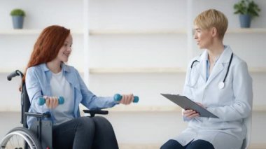 Tekerlekli sandalyedeki kızıl saçlı bir genç, bir sağlık uzmanının muayenesi sırasında küçük ağırlıkları kaldırıyor..