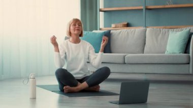 Son sınıf öğrencisi bir kadın oturma odasında yoga yapıyor, minderde bacak bacak üstüne atmış oturuyor. Odaklanmış ve sakin. Vücuduyla bağlantı kurarken farkındalık anının tadını çıkarıyor..