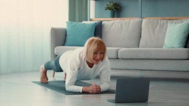 Son sınıf öğrencisi bir kadın evde antrenman yapıyor. Tahta pozisyonunu gösteriyor. Parlak bir odada çevrimiçi fitness seansını takip ederken sağlığına odaklanıyor..