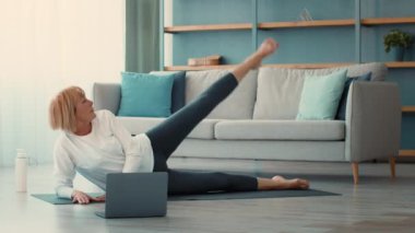 Yaşlı bir kadın oturma odasında egzersiz yapıyor, yoga minderinde bacak egzersizi yapıyor. Mekan parlak ve misafirperver, rahat bir kanepesi ve arka planda bitkileri var..