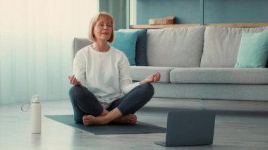 Son sınıf öğrencisi bir kadın oturma odasında bacak bacak üstüne atarak oturur, farkındalık ve egzersiz pratiği yapar. Rehberli seanslar için dizüstü bilgisayar kullanırken nefesine odaklanıyor, sakinlik yayıyor..