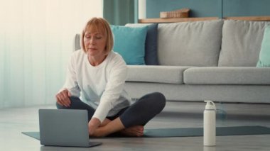 Yaşlı bir kadın yoga minderinin üzerinde oturuyor, dizüstü bilgisayarına odaklanmış bir şekilde evde online fitness rutinini takip ediyor. Yaşadığı yerde aktif ve sağlıklı kalıyor..