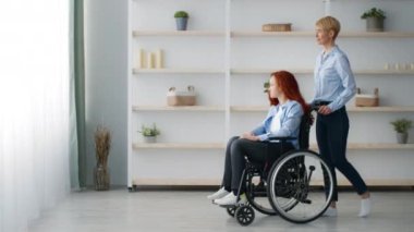 Tekerlekli sandalyedeki kızıl saçlı genç bir kız modern bir yerde rahatça oturuyor. Tekerlekli sandalye tutan bakıcı ya da anne, kopyalama alanı