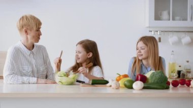 Parlak bir mutfakta, bir anne ve iki kızı yemek hazırlığı yapıyor, sebze doğruyor ve birlikte kaliteli zaman geçiriyorlar, sağlıklı yeme alışkanlıklarını ve aile bağlarını geliştiriyorlar..