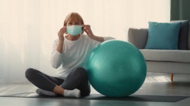 Emekli bir kadın evde spor yapıyor, güvenlik için maske takıyor. Bir sabitleme topunun yanına oturmuş, aktif kalmaya olan bağlılığını gösteriyor..