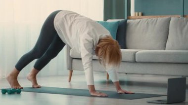 Yaşlı bir Kafkasyalı kadın evde egzersiz yapıyor, halterlerini kullanarak yoga yapıyor. Sıcak ortam, sağlık ve refah odaklı bir yaşam tarzını yansıtıyor..