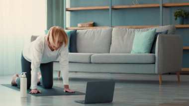 Emekli bir büyükanne, online bir çalışmanın ardından oturma odasında egzersiz yapıyor. Paspas, dizüstü bilgisayar ve su şişesi kullanırken rutinine odaklanıyor..