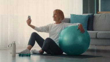 Son sınıf öğrencisi bir kadın antrenmanının tadını çıkarmak için evde spor yapıyor. Egzersiz topu kullanıyor ve aktif bir yaşam tarzı sergileyerek telefonundaki anı yakalıyor..