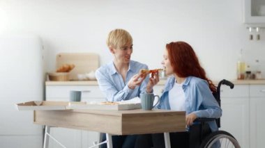 Kızıl saçlı genç bir kız tekerlekli sandalyede oturuyor, arkadaşı sıcak mutfak masasında pizzasını beslerken gülümsüyor. Parlak, davetkar bir mekanda bir anlık neşe ve dostluk paylaşıyorlar..