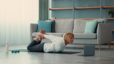 Son sınıf öğrencisi bir kadın rahat oturma odasında spor yapıyor. Paspas kullanırken esneklik ve kuvvete odaklanıyor, yanında egzersiz talimatlarını gösteren bir dizüstü bilgisayarla..