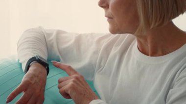 Son sınıf öğrencisi bir kadın evde spor yapıyor. Spor saatine odaklanıyor. Aydınlık oda, onun aktif ve sağlıklı kalmaktaki kararlılığını ve kararlılığını gösteriyor..