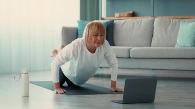 Son sınıf öğrencisi bir kadın evde yoga ve spor yapıyor. Bilgisayarında spor yapıyor. Etrafı modern bir yaşam alanıyla çevriliyken dikkatle odaklanır..