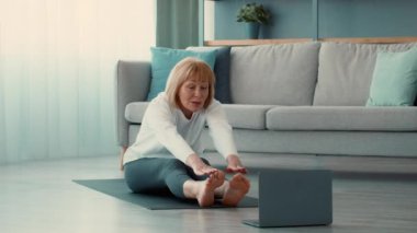 Son sınıf öğrencisi bir kadın oturma odasında yoga minderinde oturur ve online fitness dersini takip ederken gerinir. Konforlu bir ortamda egzersiz yaparken odaklanmış görünüyor..