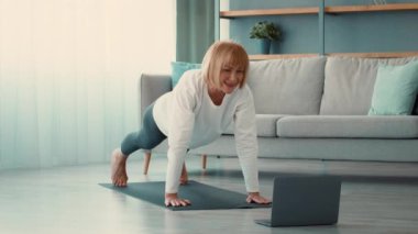 Yaşlı bir kadın, çevrimiçi bir çalışma seansından sonra evdeki yoga minderinde egzersiz yapıyor. Formuna, azmine ve rahat bir ortamda aktif bir yaşam tarzına odaklanıyor..