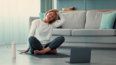 Emekli yaşlı bir kadın oturma odasında yoga rutini yapıyor, esneme ve gevşeme egzersizlerine odaklanıyor. Bir hasırın üzerinde oturuyor, online bir çalışmadan sonra..