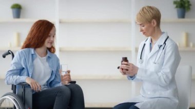 Kızıl saçlı genç bir kız tekerlekli sandalyede oturuyor ve bir sağlık uzmanıyla konuşurken gülümsüyor. Mekan rahat bir ev ortamı, rahatlık ve bakım hissi veriyor..