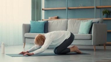 Yaşlı bir kadın evindeki paspasın üzerinde yoga pozu veriyor, spor rutinine odaklanıyor. Parlak ve rahat oturma odası egzersiz için huzurlu bir ortam yaratıyor..