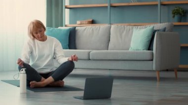 Kafkasyalı emekli bir kadın, oturma odasındaki yoga minderinde meditasyon yapıyor. Çevrimiçi bir dersi takip ederken bir anlık huzurun tadını çıkarıyor..