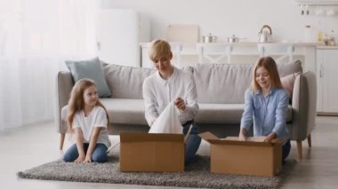 Bir aile keyifli bir şekilde oturma odasındaki kutuları açıyor. Anne giysi taşırken, kız ve anne tableti kontrol ediyor..
