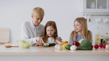 Bir ebeveyn iki küçük çocuğa taze sebzelerle dolu modern bir mutfakta sağlıklı bir yemek hazırlamayı öğretiyor. Kahkaha ve neşe yemek yaparken boşluğu doldurur..