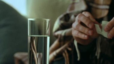 30 'lu yaşlarında bir adam kendini evinde bir battaniyeye sarmış, hasta ve izole bir halde görünüyor. Bir bardak suyla otururken ilaç hazırlıyor, açıkça soğuk algınlığı veya grip ile uğraşıyor..