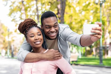 Neşeli siyahi adam tekerlekli sandalyedeki engelli kız arkadaşına sarılıyor sonbahar parkında akıllı telefondan selfie çekiyor. Engelli çift dışarıda dolaşırken kendi fotoğraflarını çekiyorlar.