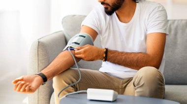 Evdeki kanepede oturan genç Hintli adam kan basıncını ölçüyor. Orta Doğulu beyaz tişörtlü adam modern tonometre kullanıyor. Stres kaynaklı hipertansiyon yaşıyor.