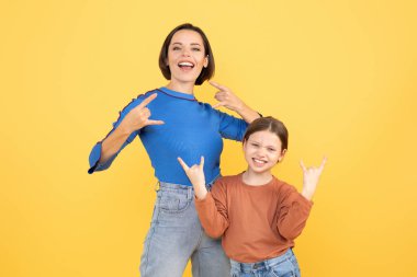 Neşeli bir anne ve küçük kızı birlikte sevgi dolu bir anı paylaşıyor, neşeli jestler yapıyor ve birbirlerine olan sevgilerini canlı sarı bir zemine yansıtıyorlar..