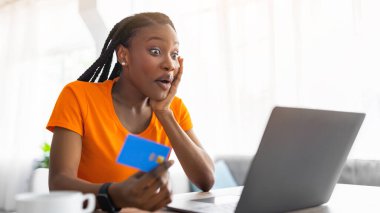 Duygusal Afro kadın dizüstü bilgisayara bakıyor, elinde kredi kartı var, web mağazasında büyük bir satıştan şok olmuş. Evde otururken internet üzerinden mal ve hizmetlere para ödeyen siyahi kadın şaşırdı.