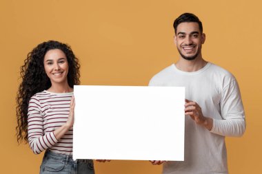 Ad Mockup. Mutlu genç Arap eşler, ellerinde beyaz boş levhalarla, gülümseyen orta doğulu çift, sarı stüdyonun arkasında dururken tasarımınız için fotokopi alanı gösteriyor.