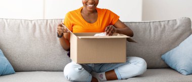 Tam boy, karton paketi açan pozitif siyahi kadın, eve çevrimiçi teslimat alıyor. Genç bayan müşteri, müşteri hizmetlerinden memnun, web sitesi siparişini açıyor.