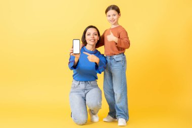 Bir anne ve küçük kızı, canlı sarı bir arkaplan karşısında akıllı bir telefonla poz verirken şefkat gösterirler. Her ikisi de neşeli gülücükler sergiliyor ve neşeli pozlar veriyor..