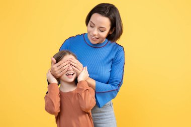 Sevgi dolu bir anne, kızının gözlerini neşe içinde kapatırken kız sevinçle gülümsüyor. Parlak sarı bir arkaplanda izole edilmişler, yakın bağlarını ve mutluluklarını sergiliyorlar..