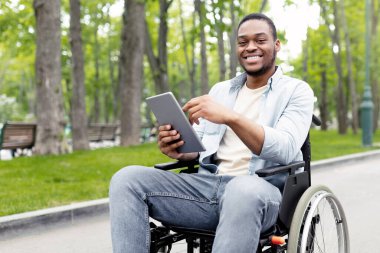 Şehir parkında uzaktan haberleşme için dizüstü bilgisayar kullanan tekerlekli sandalyedeki sakat genç adamın portresi. Özürlü mutlu siyah adam online toplantı yapıyor, dışarıda arkadaşlarıyla sohbet ediyor.