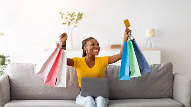 Kanepede dizüstü bilgisayarı ve kredi kartıyla oturan neşeli siyahi kadın, online alışveriş yapıyor, evden satın alıyor. Elektronik mağaza servisini kullanarak hediye çantaları taşıyan heyecanlı bayan müşteri