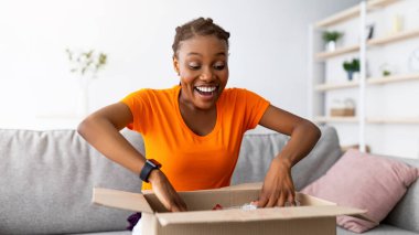 Duygusal siyahi kadın müşteri uzun zamandır beklenen paketi alıyor, karton kutuyu evde açıyor, büyük alışverişlerle tatmin oluyor. Çevrimiçi alışveriş ve dağıtım kavramı