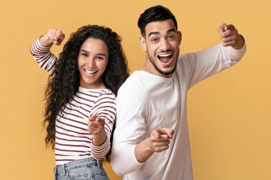 Yakaladım seni. Neşeli orta doğulu çift kamerayı parmaklarıyla işaret ediyor, neşeli heyecanlı Arap erkek ve kadın birini işaret ediyor ve sarı arka planda dururken birlikte eğleniyorlar, boş alan.
