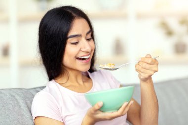 Dengeli beslenme konsepti. Güzel Hintli bayan evdeki rahat kanepede sütle lezzetli mısır gevreği yiyor. Sağlıklı yaşam tarzını sürdüren güzel bir doğu kadını, evde sağlıklı yemekler yiyor.