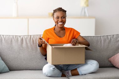 Karton paketi açan neşeli siyahi bir kadın, bacak bacak bacak üstüne atmış koltukta oturuyor, içerde kameraya gülümsüyor. Genç kadın tüketici çevrimiçi market siparişini açıyor, teslimat hizmetinden memnun.
