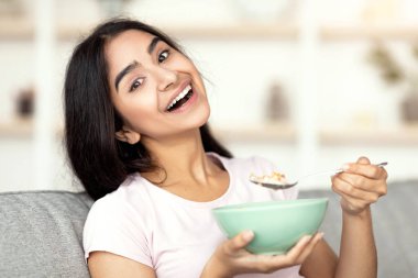 Evdeki rahat koltukta gevrek ve süt yiyen neşeli Hintli kadın kameraya gülümsüyor. Sağlıklı yaşam tarzını sürdüren, sağlıklı yemek yiyen güzel bir doğu kadını. Dengeli beslenme kavramı