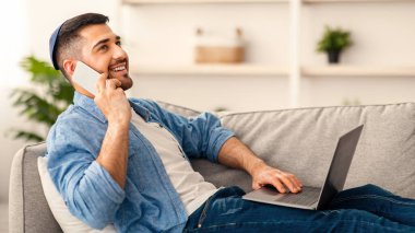 Uzak Çalışma ve Telekomünikasyon. Cep telefonuyla konuşan, bilgisayarını uzaktan kullanan, oturma odasındaki kanepede oturan, boş alan seçici odaklı mutlu bir Yahudi.