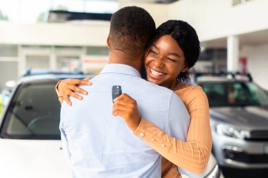 En iyi hediye. Mutlu Siyah Kadın Araba Anahtarı Tutuyor ve Satış Merkezi 'nden yeni bir araba aldıktan sonra kocasını kucaklıyor, Neşeli Afrikalı Amerikalı Kadın Hediyelik, Kapalı Çekim,