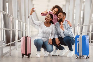 Üç kişilik mutlu siyah aile selfie çekiyor, havaalanında bavulla poz veriyor, akıllı telefona sarılıyor ve neşeyle gülümsüyor zafer işareti gösteren bir adam, el kol hareketi yapan bir kız.